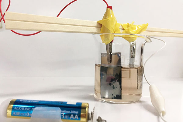 図2　ポリアニリンの電界重合。電池の＋極につながっているステンレス板には、緑色の膜のような物質が付着している。これがポリアニリン。（溶液は少しアニリンが酸化したため、褐色ですが、新しいアニリンであれば無色透明です。またポリアニリンは時間をかけると全体につきます。部分的についているだけでも、充電・放電はできます。）